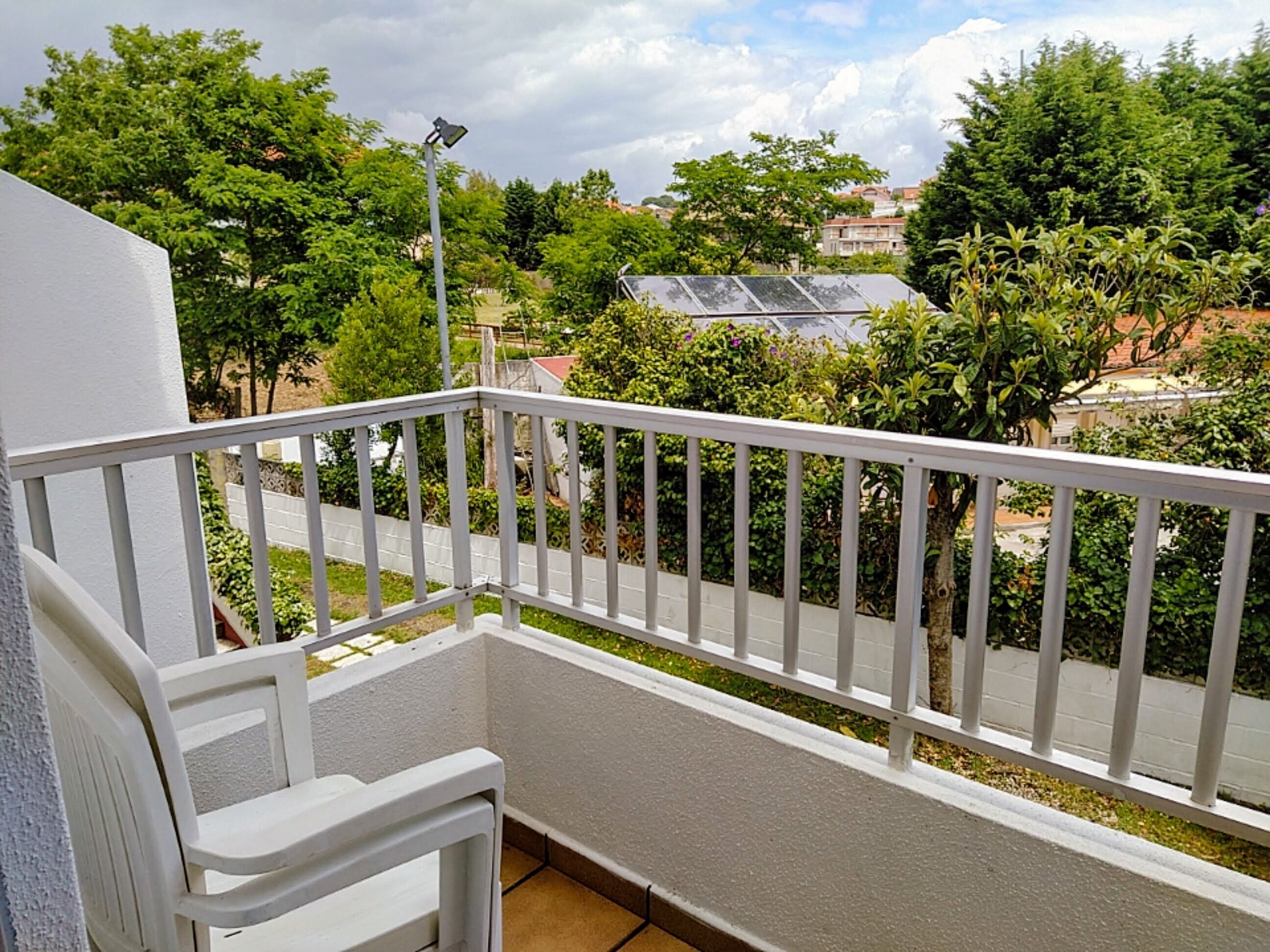 Ein Balkon mit weißer Brüstung, ein kleiner weißer Stuhl und Blick auf eine Straße mit Grünflächen sowie ein Gebäude mit Solaranlage.