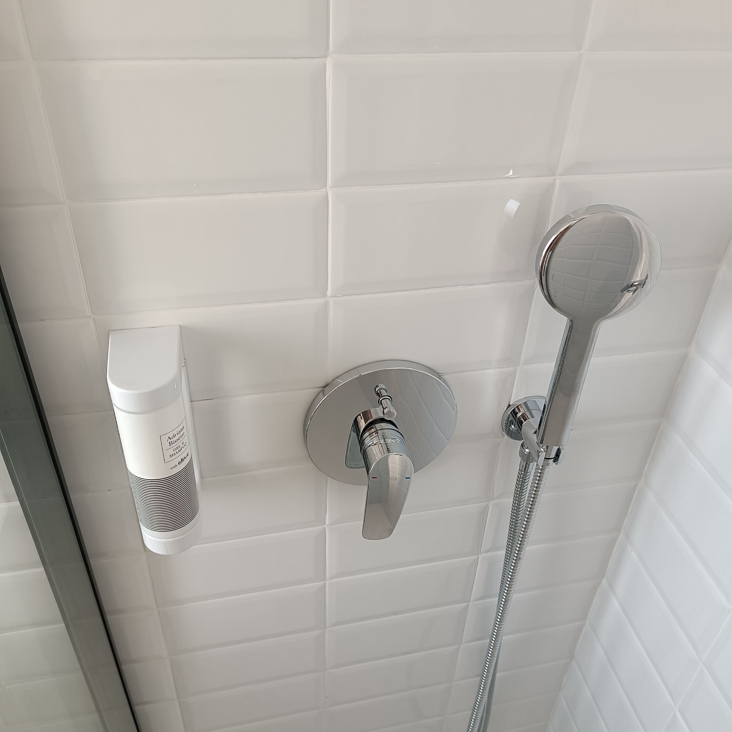A bathroom with white tiled walls, a handheld showerhead, and a bottle of shampoo.