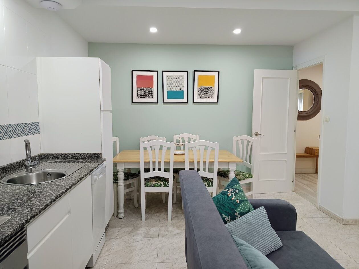 Una cocina compacta con comedor, armarios blancos, encimera de granito y una mesa con sillas. Hay tres obras de arte enmarcadas en la pared sobre la mesa.