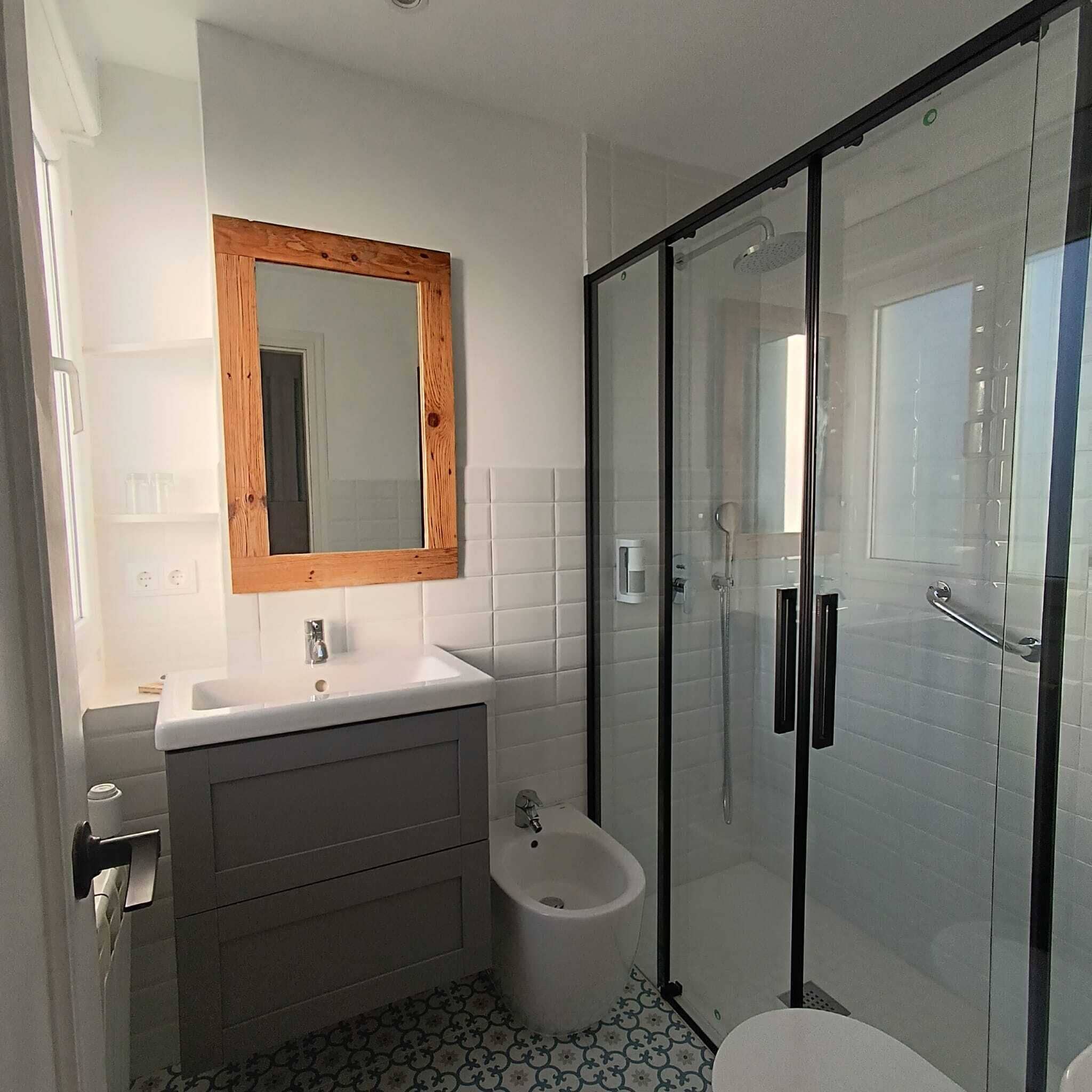Bany modern amb un rentamans blanc, un armari de tocador de fusta, una cabina de dutxa de vidre i rajoles de terra amb un disseny original.