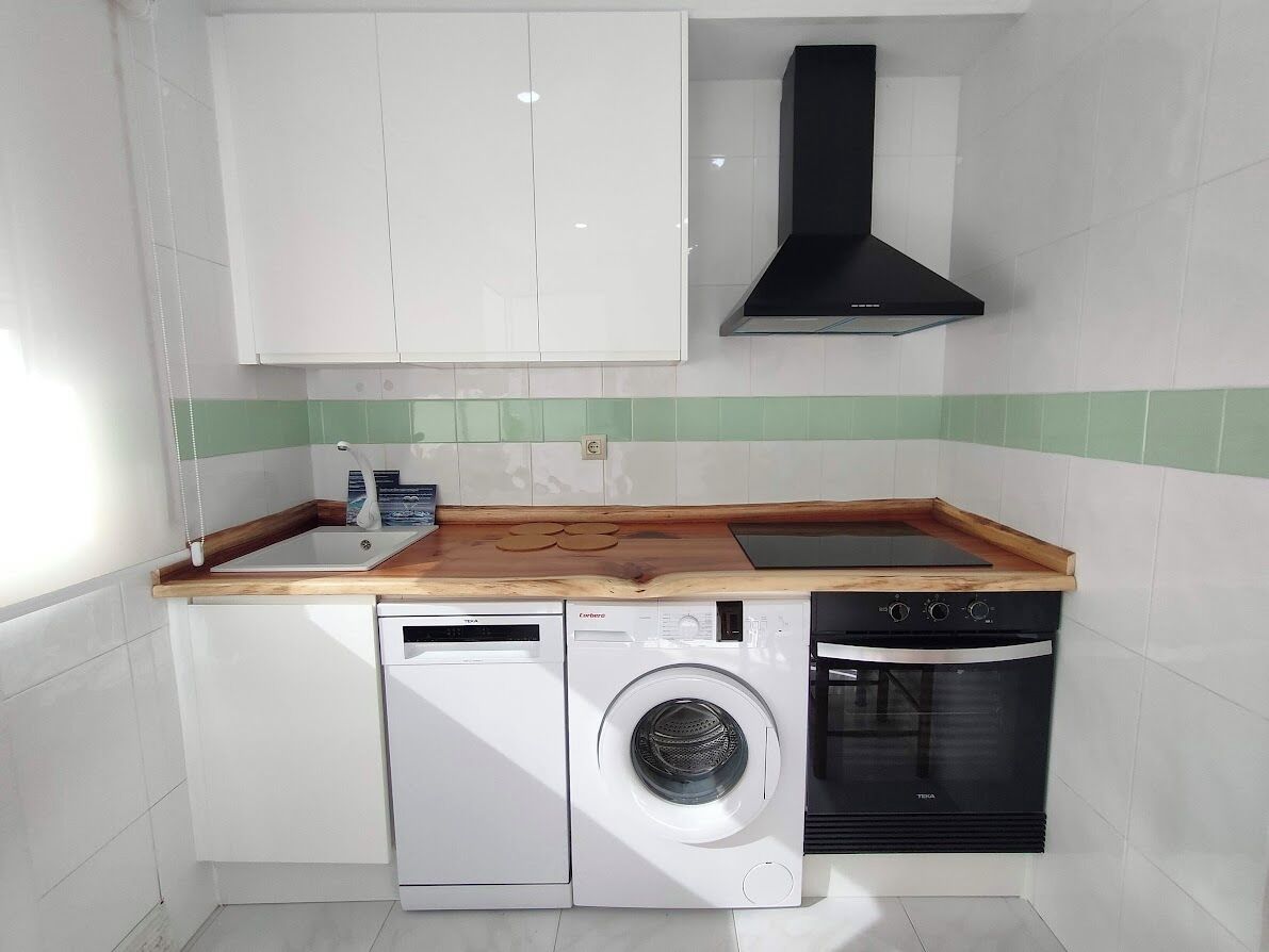 A compact kitchen with white cabinets, a black range hood, a dishwasher, a washing machine, and a built-in oven.