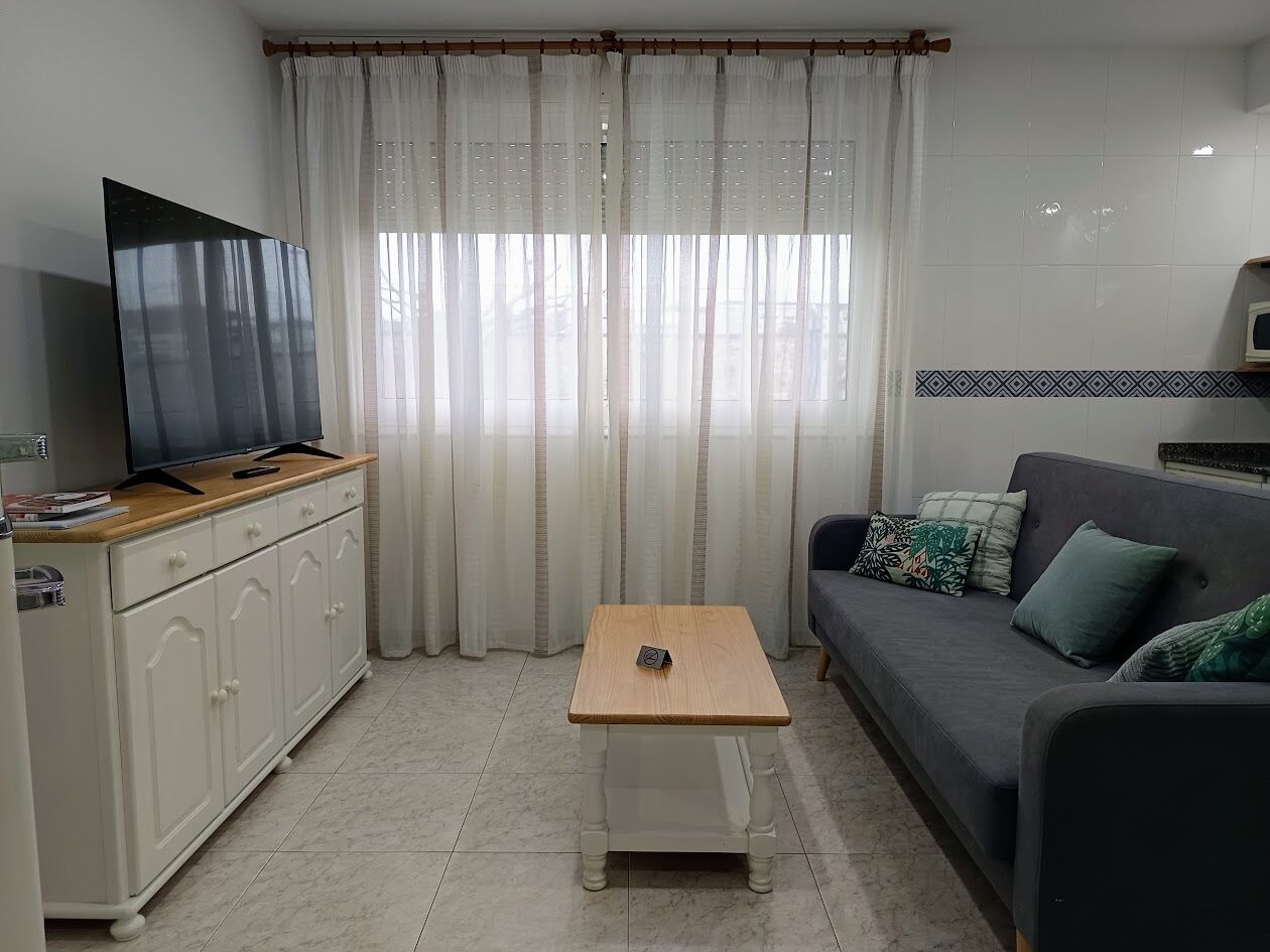 A living room with a flat-screen TV on a wooden stand, a grey sofa with patterned cushions, a small wooden table, and a white cabinet.