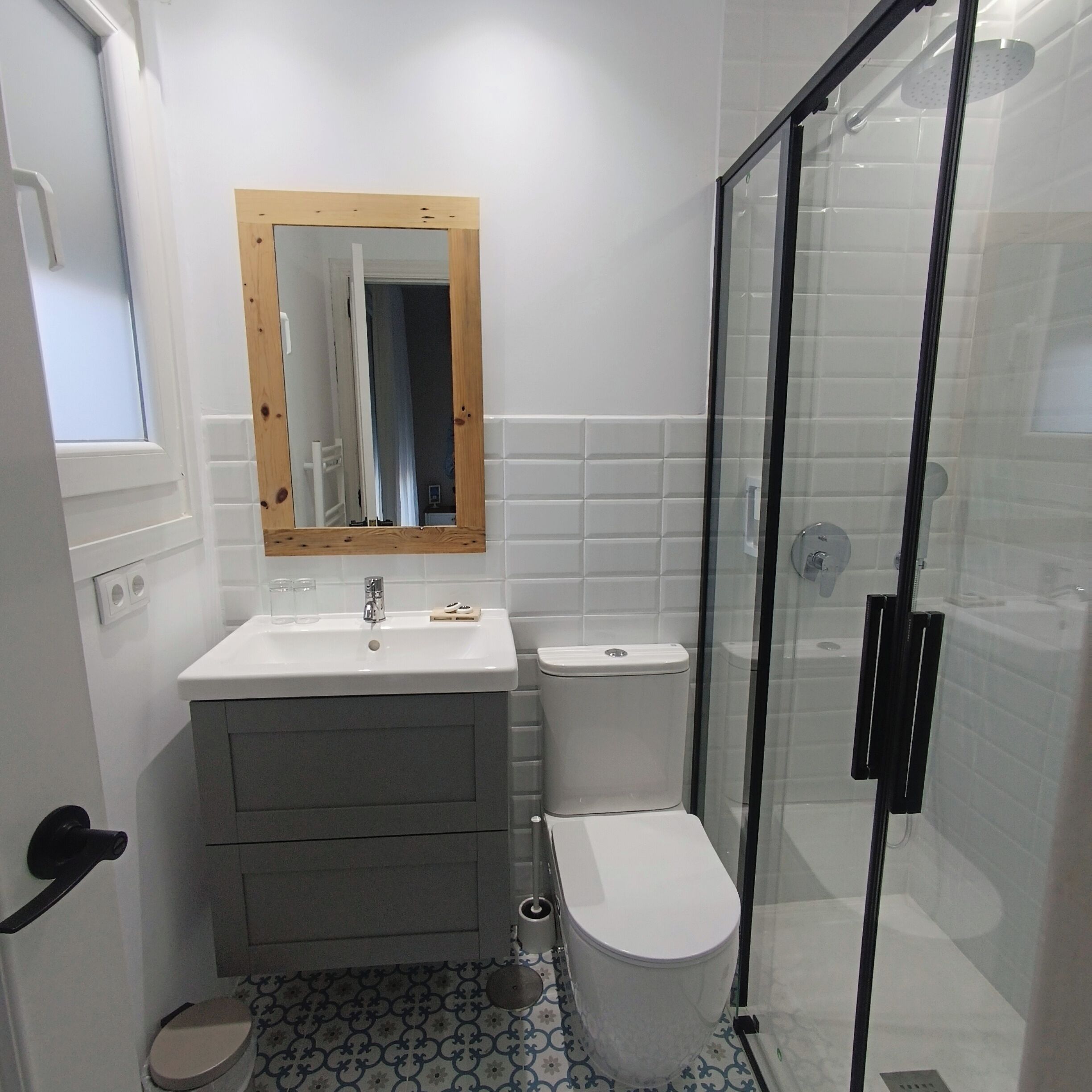 Un baño con un lavamanos blanco, un espejo con marco de madera, un inodoro y una cabina de ducha con paredes de vidrio.