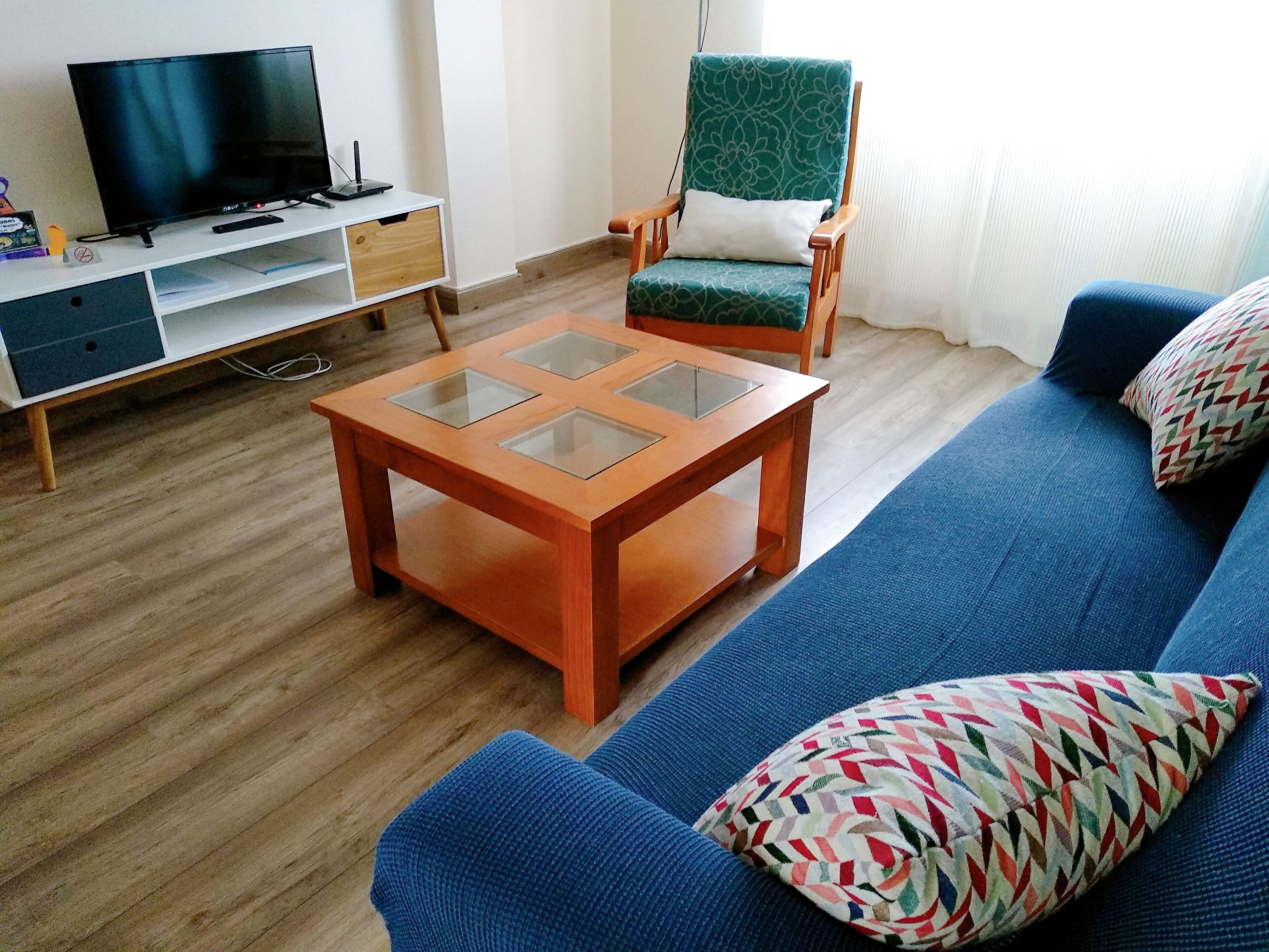A living room with a blue sofa, a wooden coffee table with glass panels, a green patterned armchair, and a TV on a white stand.