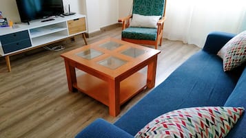A living room with a blue sofa, a wooden coffee table with glass panels, a green patterned armchair, and a TV on a white stand.