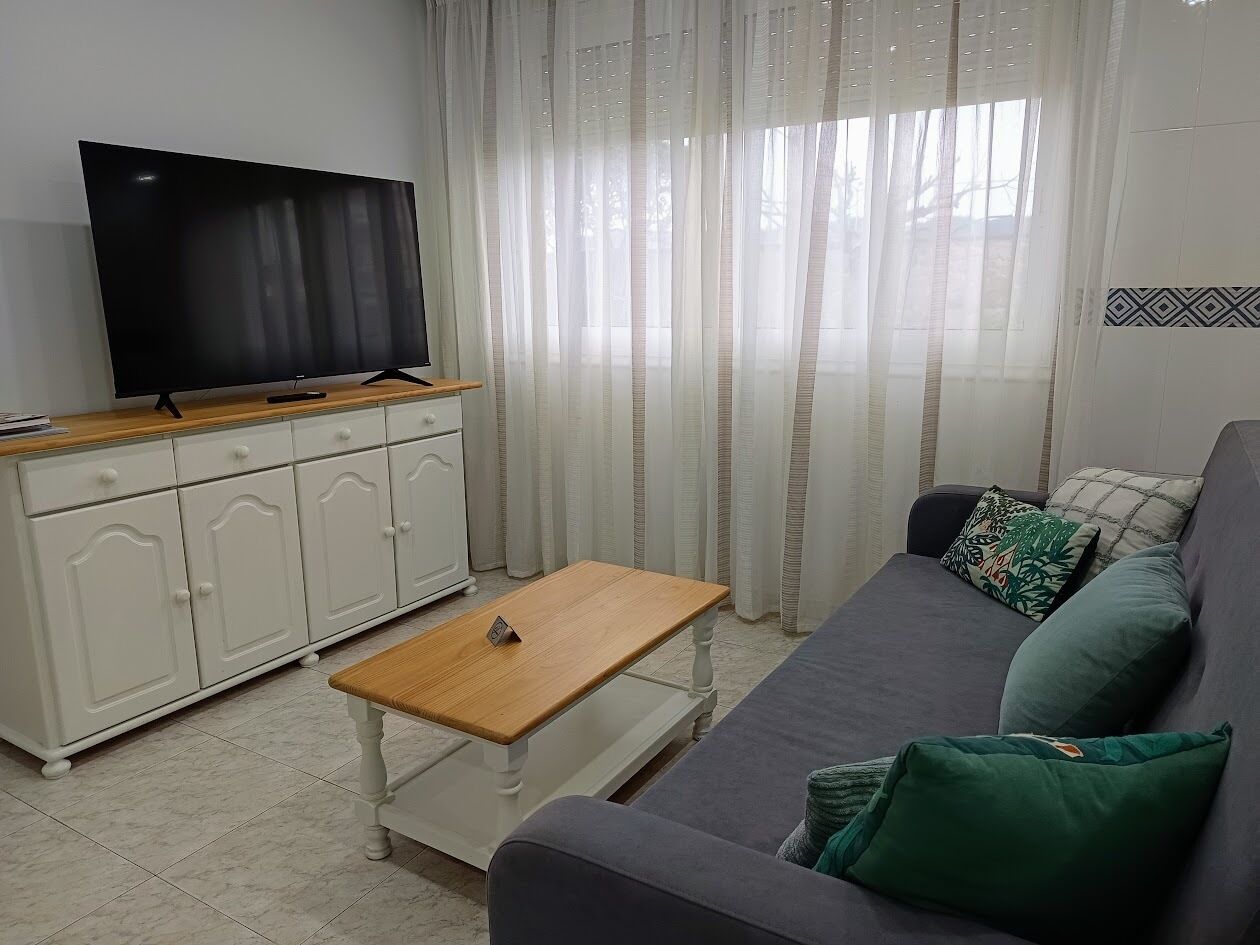 Una sala de estar con un televisor sobre un mueble de madera, un armario blanco, un sofá gris con cojines verdes y una mesa de centro de madera.