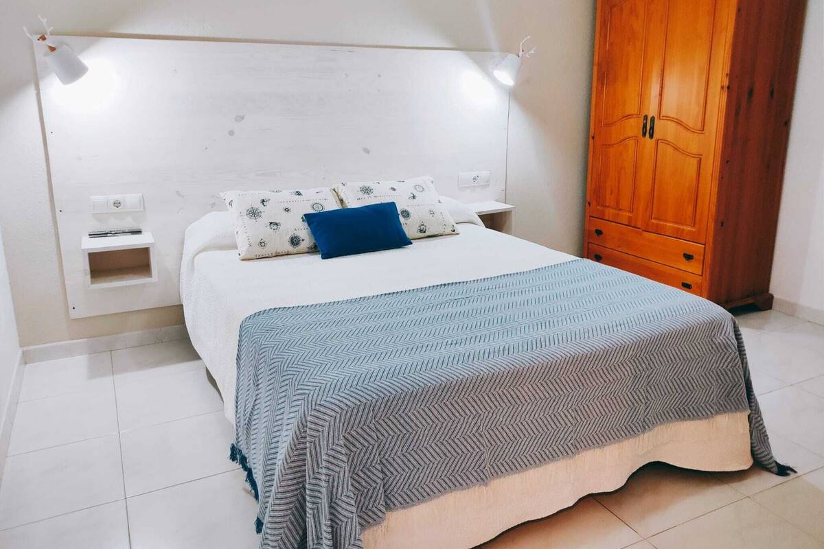 A bedroom with a bed, a nightstand, a wooden wardrobe, and a wall-mounted light fixture.