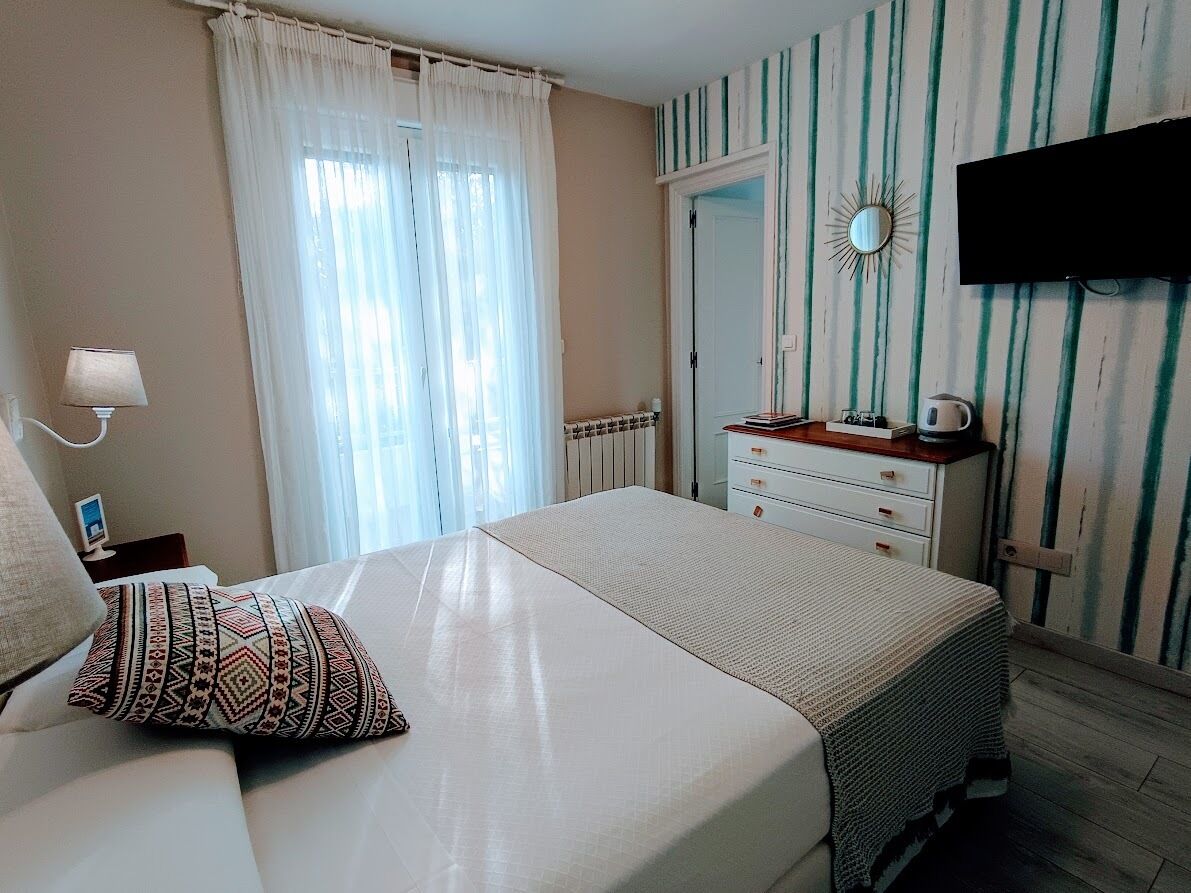 A bedroom with a bed, a bedside table with a lamp, a dresser, a mirror, and a television mounted on the wall.