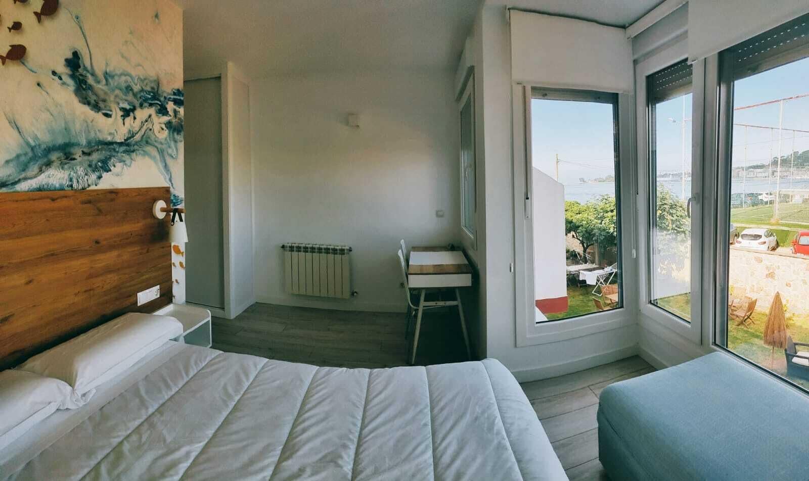 A bedroom with a bed, a wooden headboard, a radiator, a small table, and a view of the outdoors through a large window.