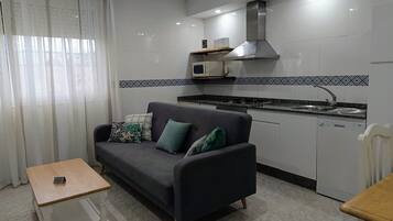 Un ambiente compatto con divano, un piccolo tavolo, angolo cottura con forno a microonde ed elettrodomestici.