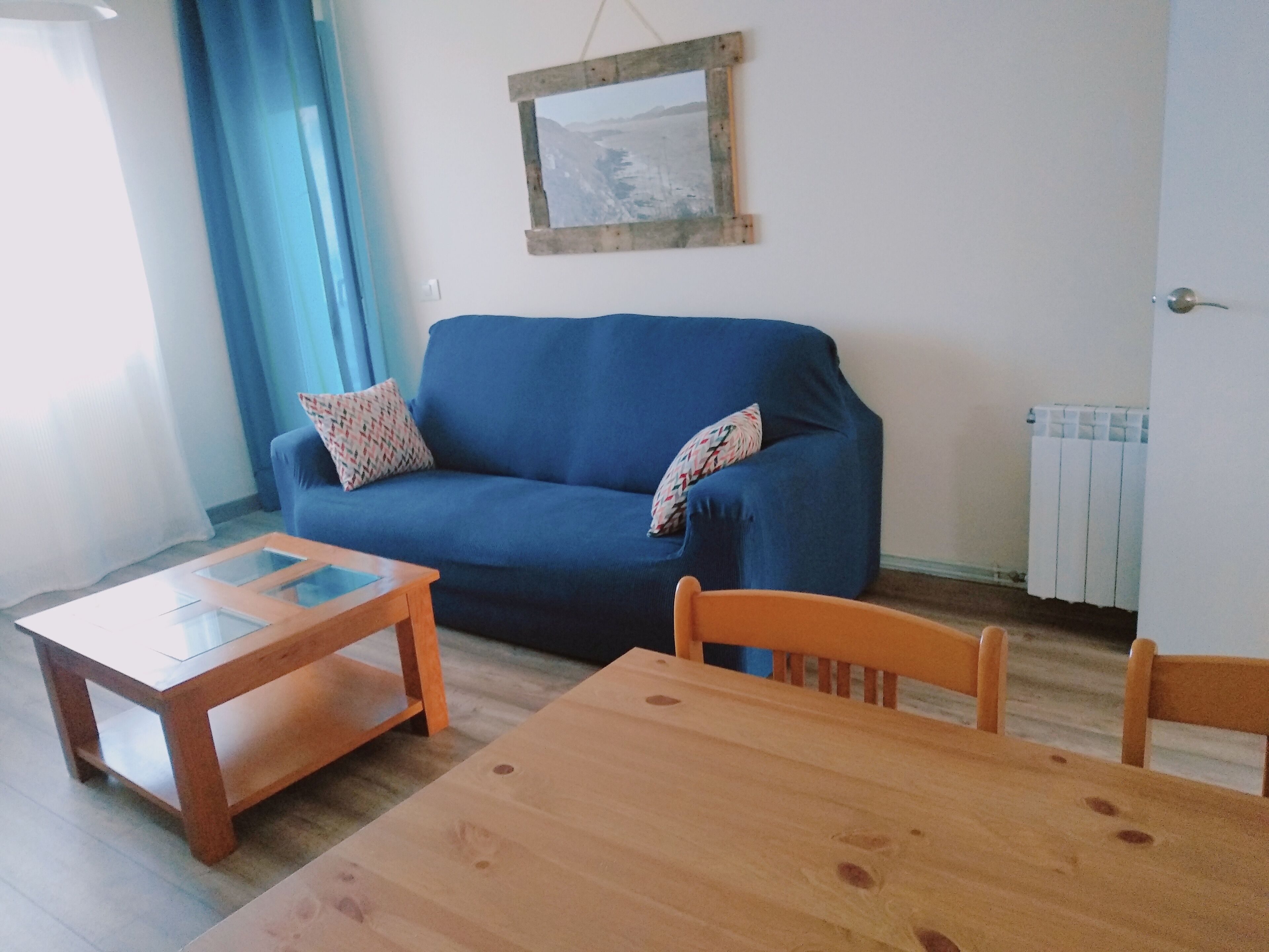 A living room with a blue sofa, two patterned pillows, a wooden coffee table, and a wooden dining table.