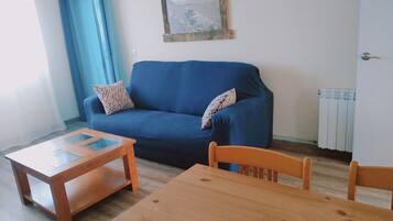Una sala de estar con un sofá azul, dos almohadas estampadas, una mesa de centro de madera y una mesa de comedor de madera.