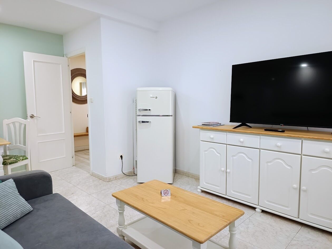 Un espace de vie compact comprenant un canapé, une petite table en bois, un meuble blanc avec un téléviseur et un réfrigérateur blanc.
