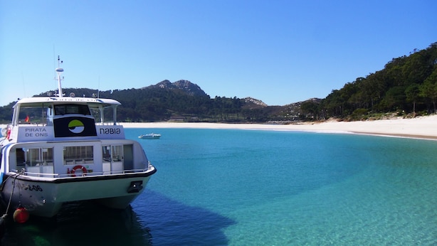 A boat named 'PIRATA DE ONS' docked near a clear blue beach with white sand and a forested hill in the background. - Hotel Rodeiramar (Cangas)