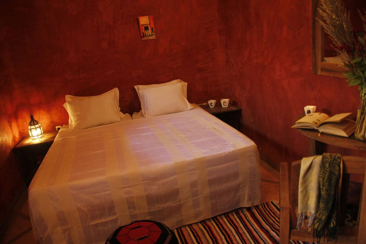 A neatly made bed with a white bedspread and pillows, a bedside table with a lamp, a small nightstand with a phone and a cup, and a chair with a blanket.