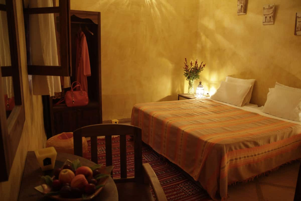 A room with a bed, a nightstand with a lamp, a chair, a table with fruit, and a hanging bag.