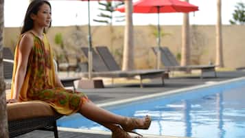 A person sitting by a poolside with red umbrellas and lounge chairs in the background.