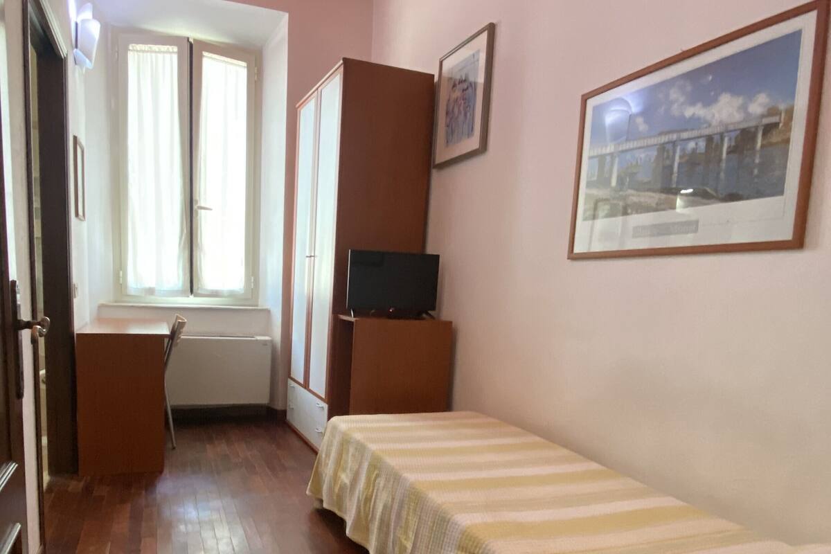 A small, sparsely furnished room with a wooden floor, a single bed, a desk, a chair, a television on a stand, and a window with curtains.