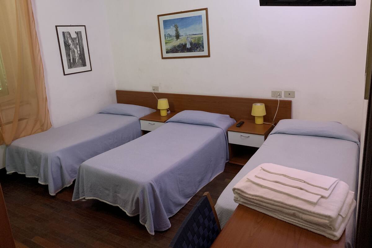 A hotel room with two single beds, a wooden nightstand with lamps, a small table with a stack of towels, and two framed pictures on the wall.