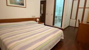 A hotel room with a bed, a nightstand, a lamp, a mirror, a wardrobe, and a bathroom visible through an open door.