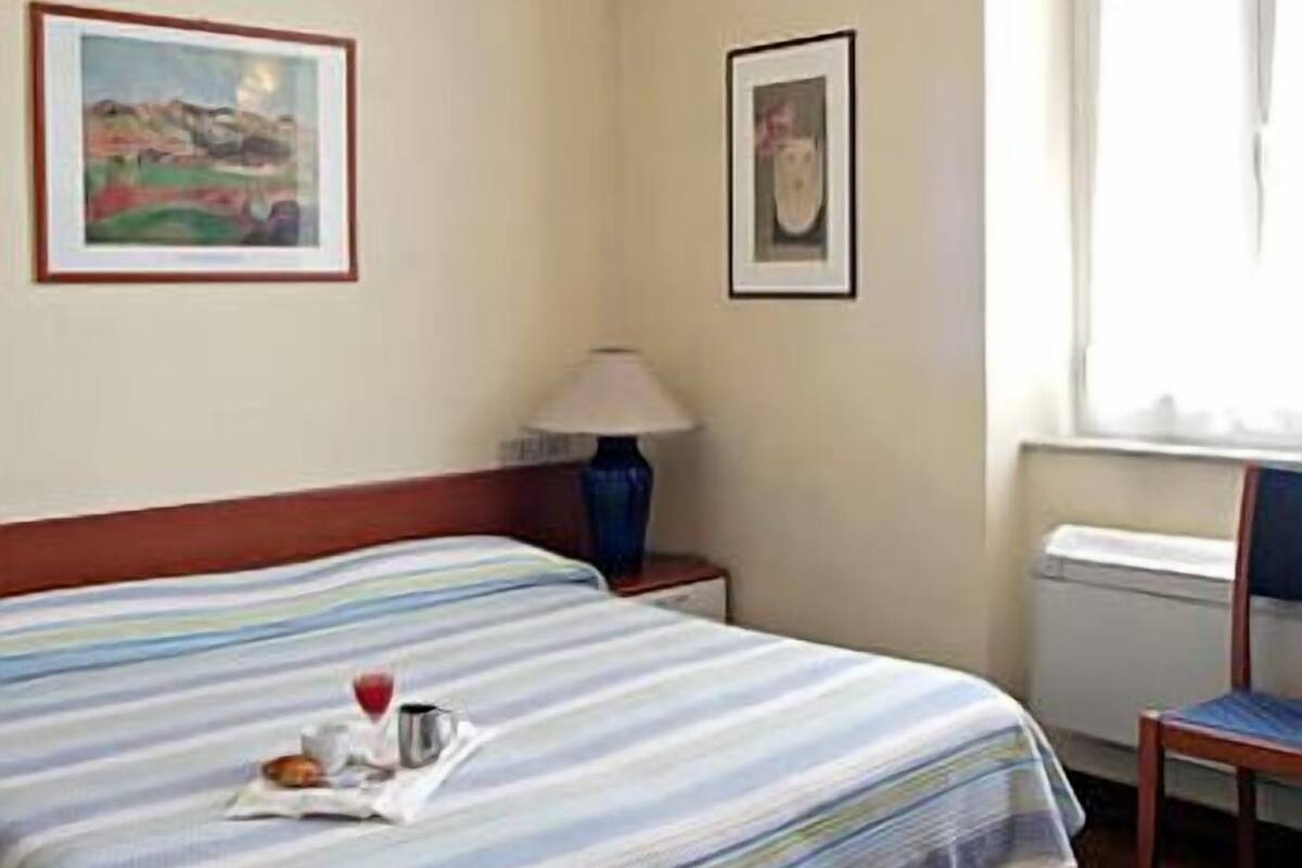 A hotel room with a bed, a lamp, a chair, and two framed pictures on the wall.