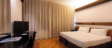 A hotel room with a bed, a desk with a television, and a large window with curtains.
