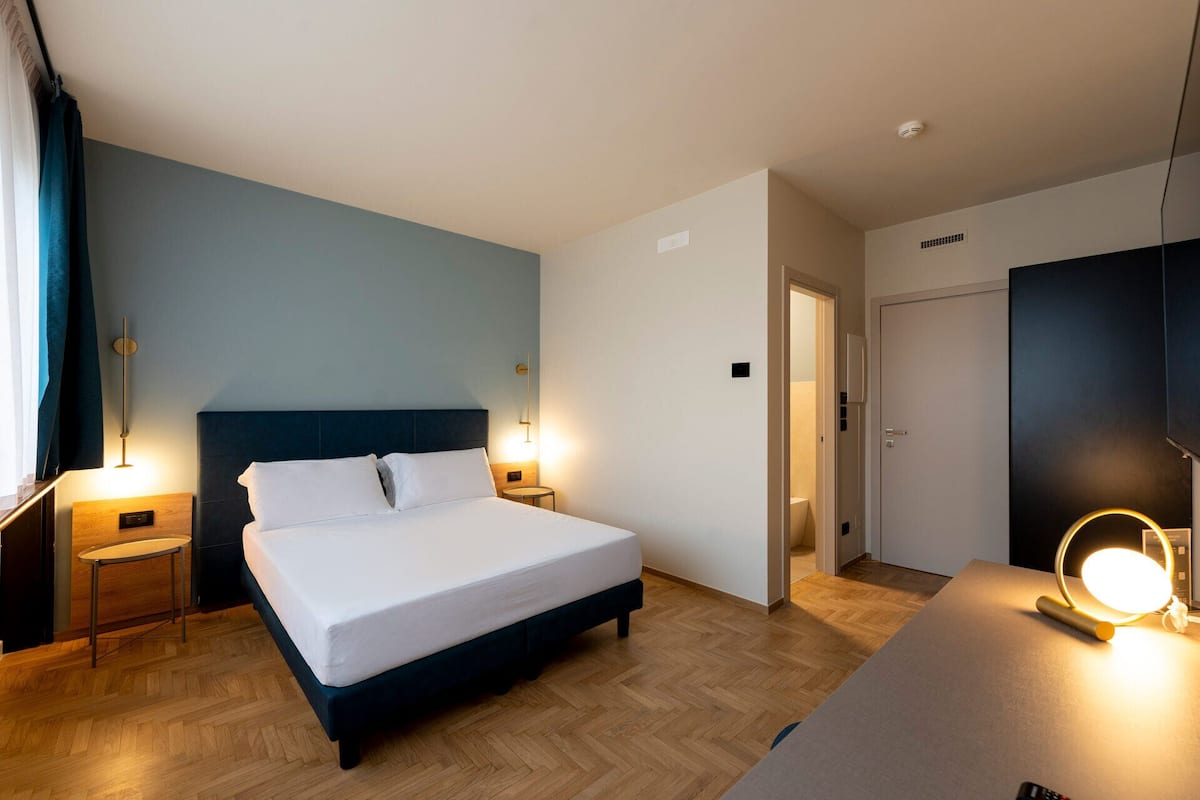 A hotel room with a bed, bedside tables, a desk with a lamp, and a wardrobe.