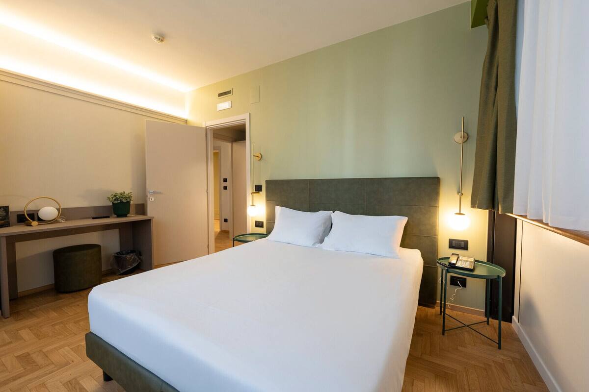 A hotel room with a large bed, a desk with a chair, a small table with a lamp, and a mirror on the wall.