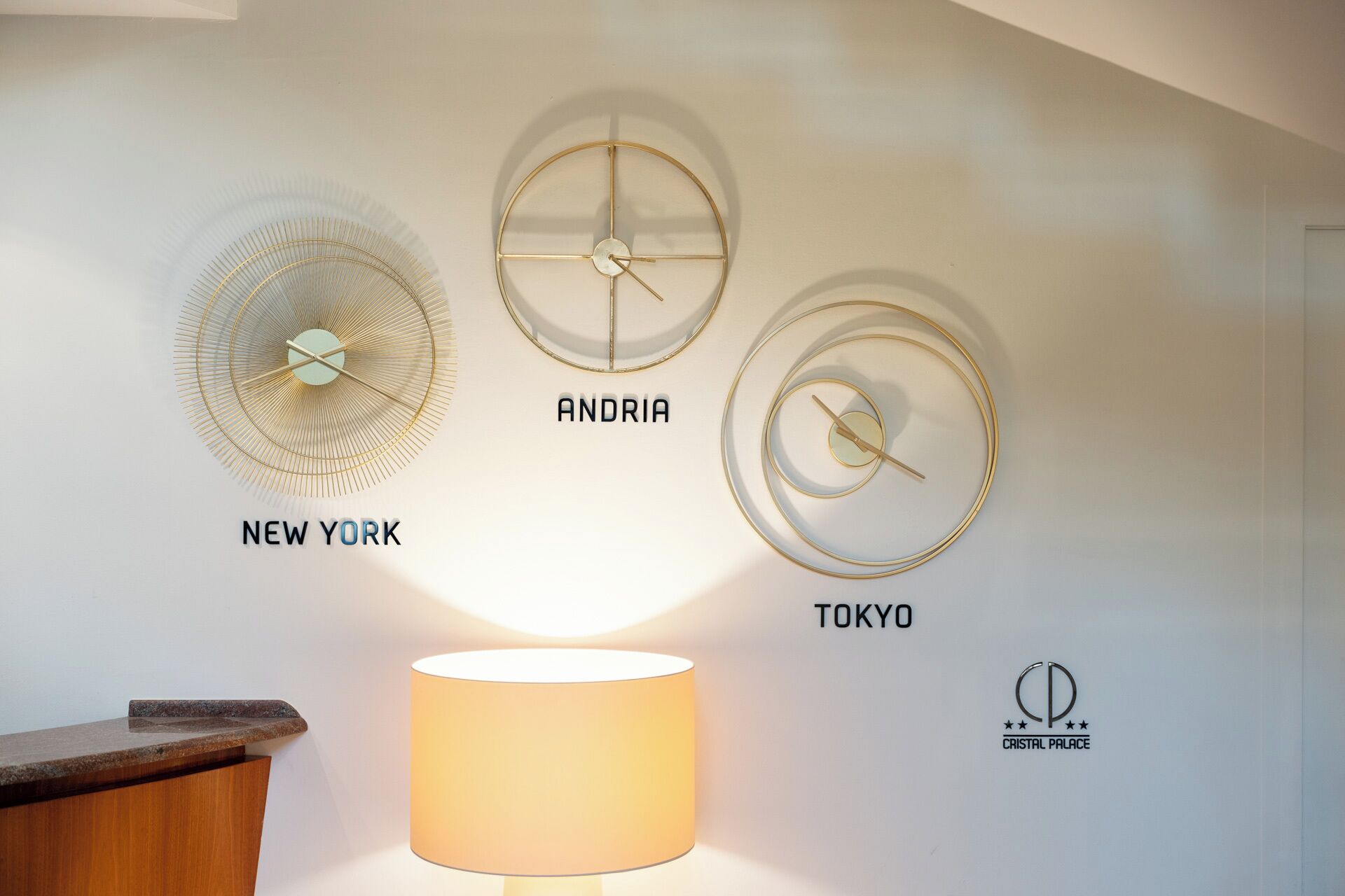 Un muro con tre orologi da parete circolari etichettati 'NEW YORK', 'ANDRIA' e 'TOKYO', e una lampada sotto.