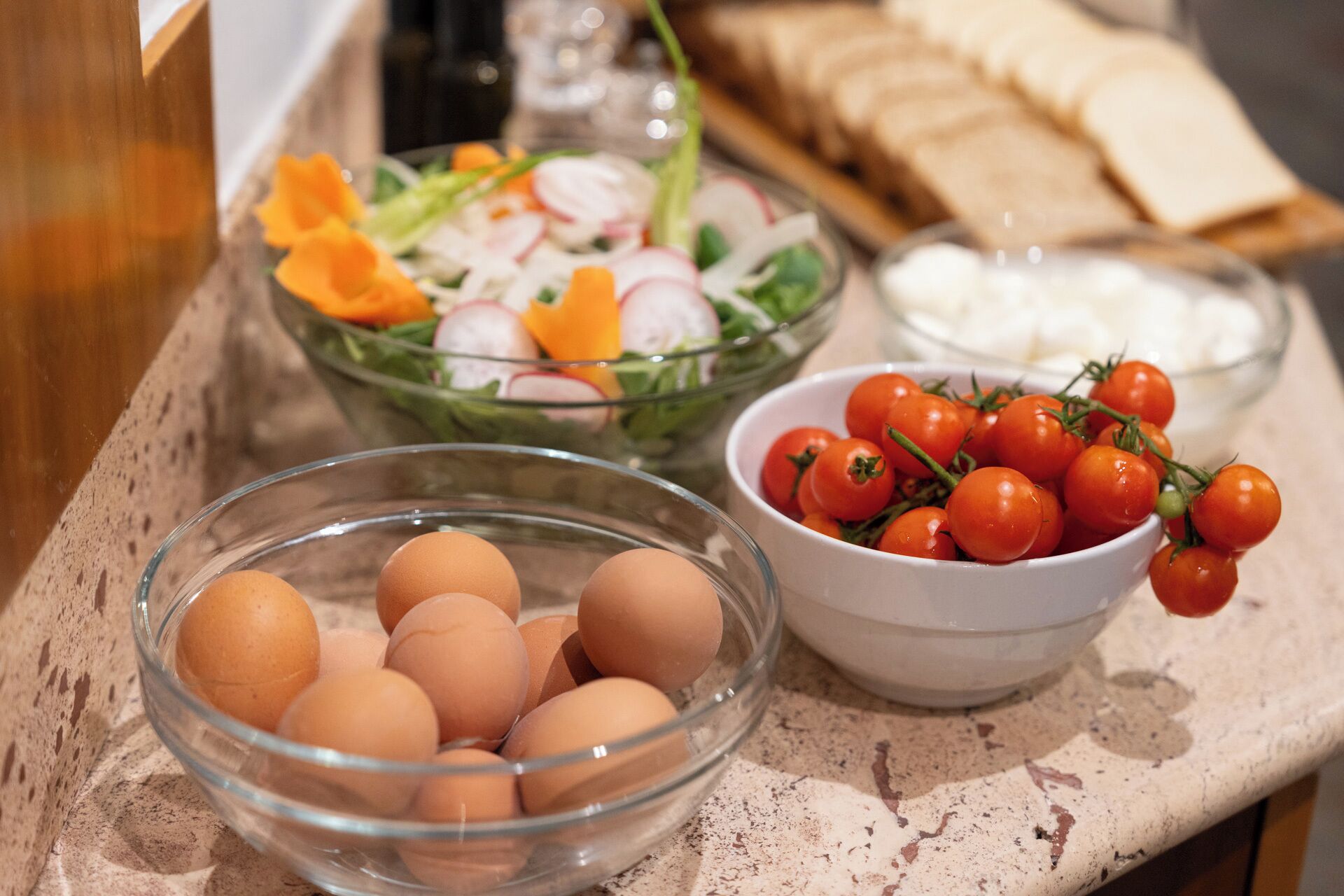 A kitchen counter with various food items including eggs, cherry tomatoes, a salad, and sliced bread.