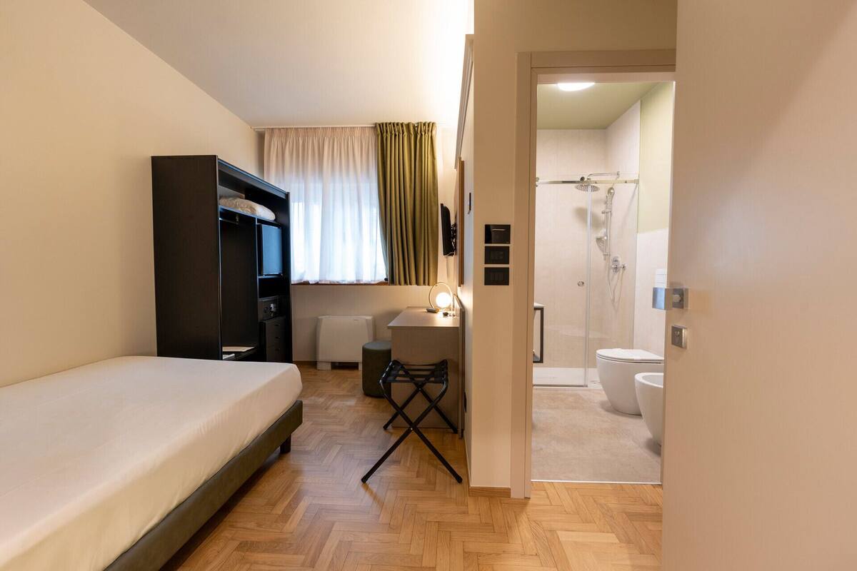 A compact hotel room with a single bed, a desk with a chair, a wardrobe, a bathroom with a shower, toilet, and sink.