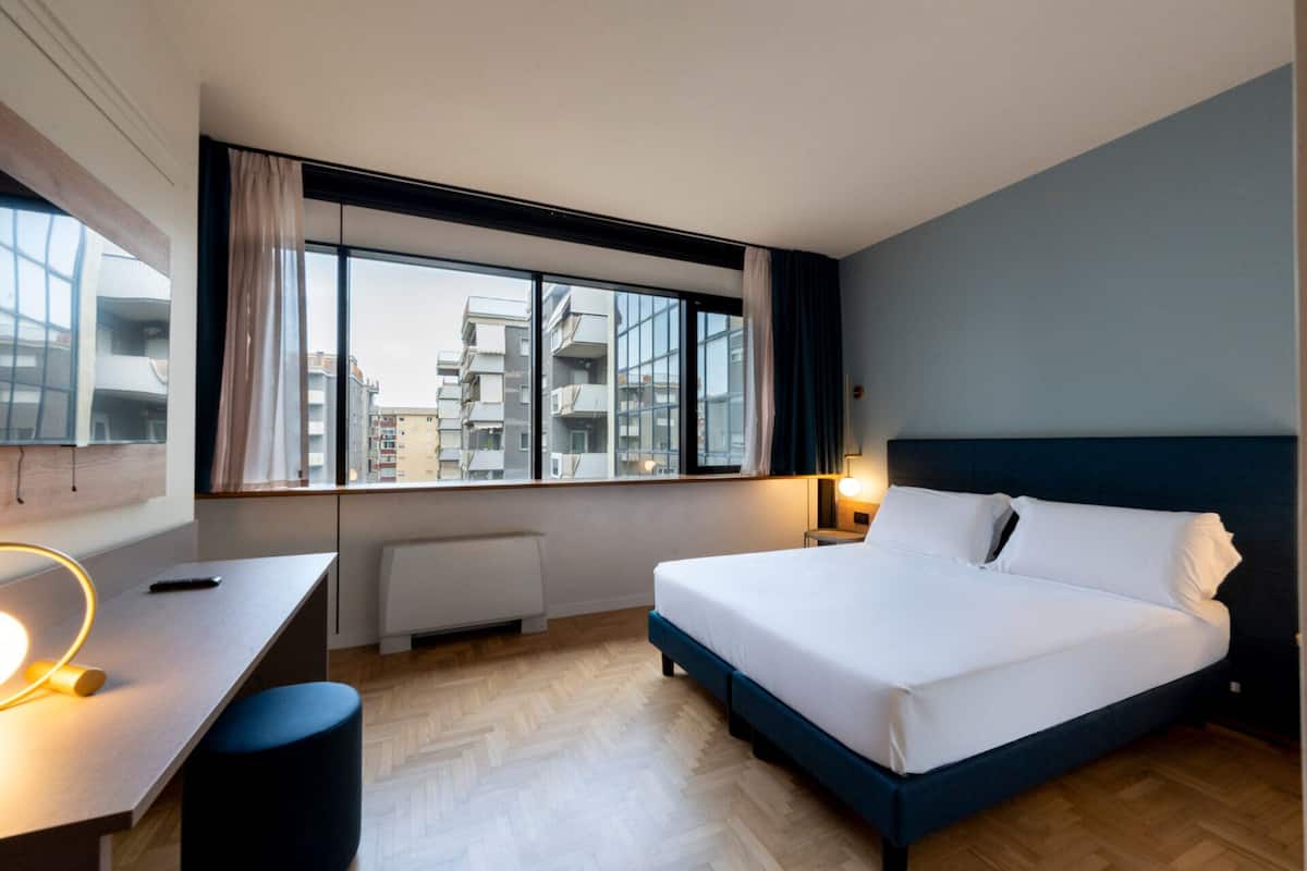 A hotel room with a bed, a desk, a chair, a window with a view of buildings, and a radiator.