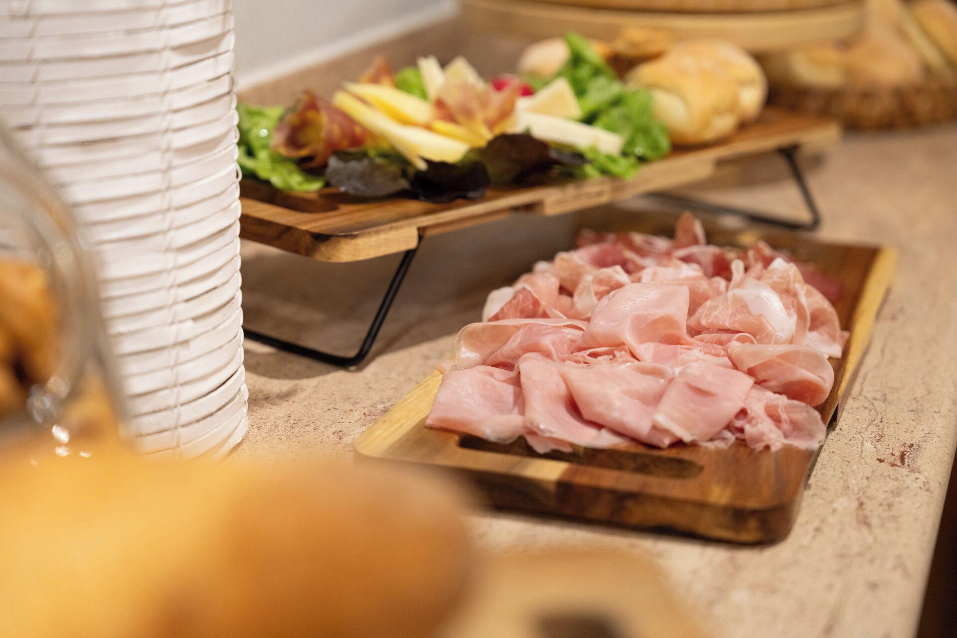 Una bandeja de madera con jamón en rodajas, un tarro de cristal y una tabla de madera con diversos alimentos.
