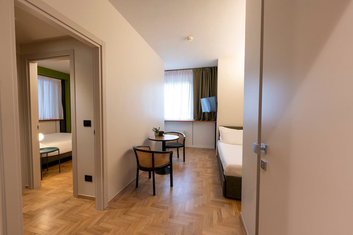 A hotel room with a bed, a small table with chairs, a TV on a stand, and a window with curtains.