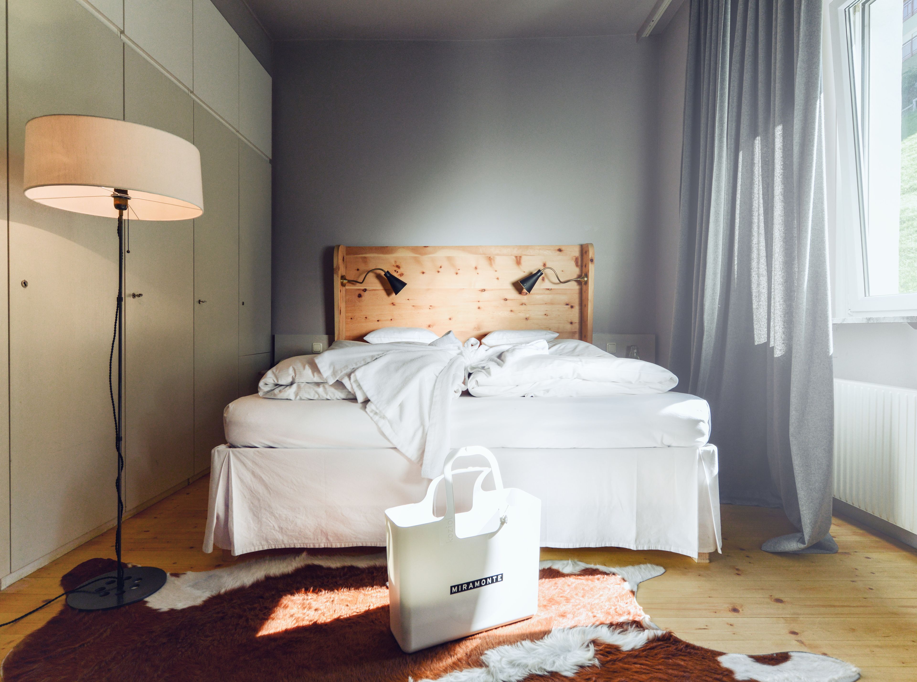 A bedroom with a wooden headboard, white bedding, a standing lamp, and a fur rug.
