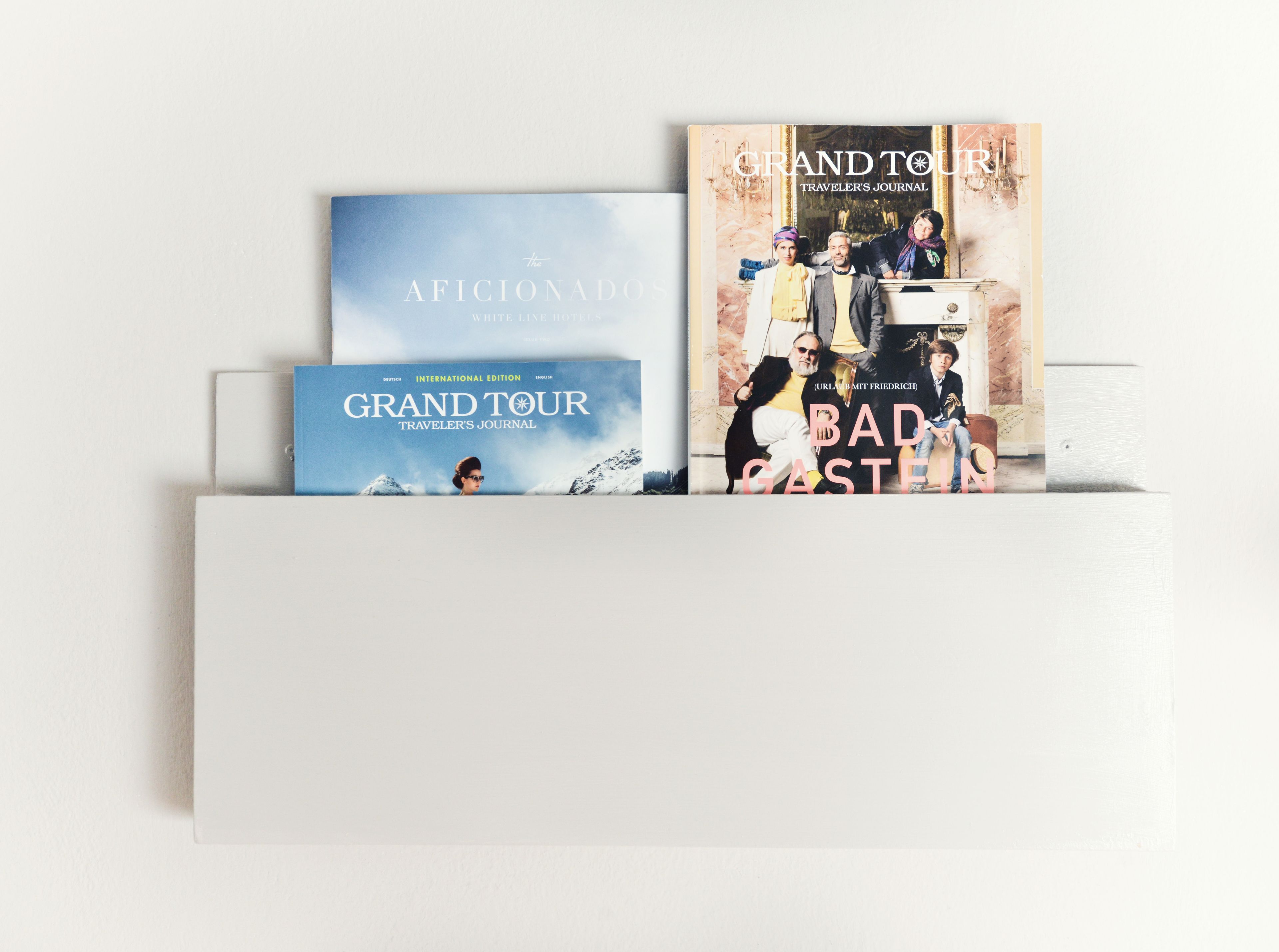 A white shelf with three books: 'The Aficionado' by White Line Hotel, 'Grand Tour' by International Edition, and 'Bad Gastein' by URL.comit Friedrich.