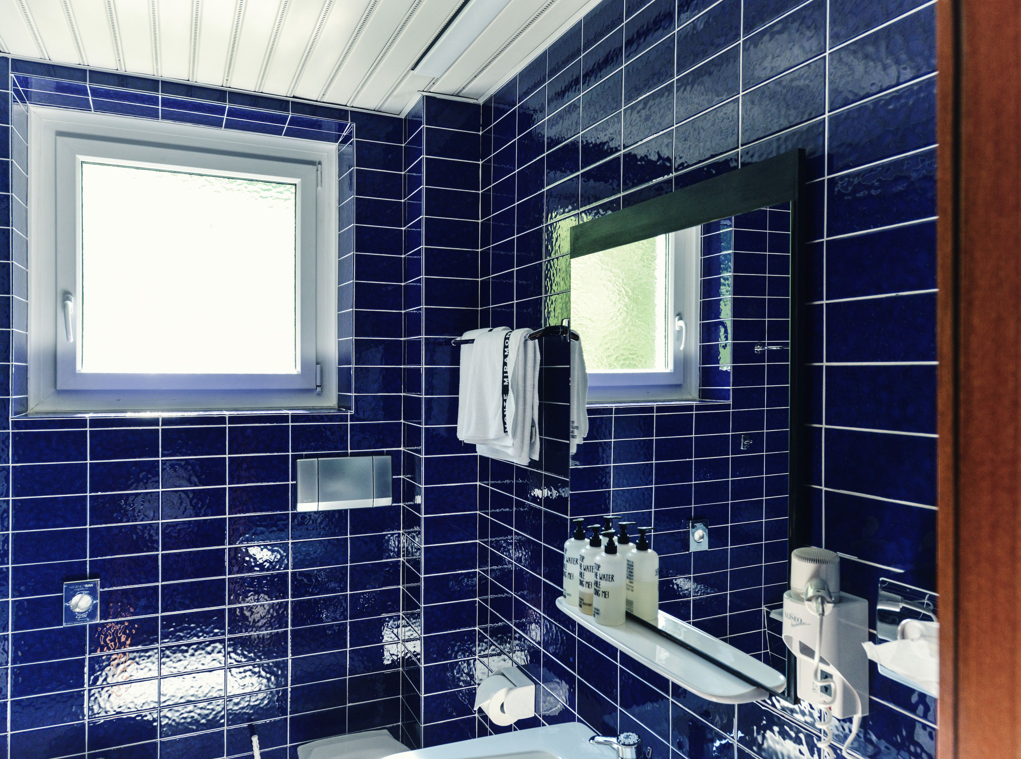 A bathroom with blue tiles, a window, a towel rack, and a shelf with toiletries.