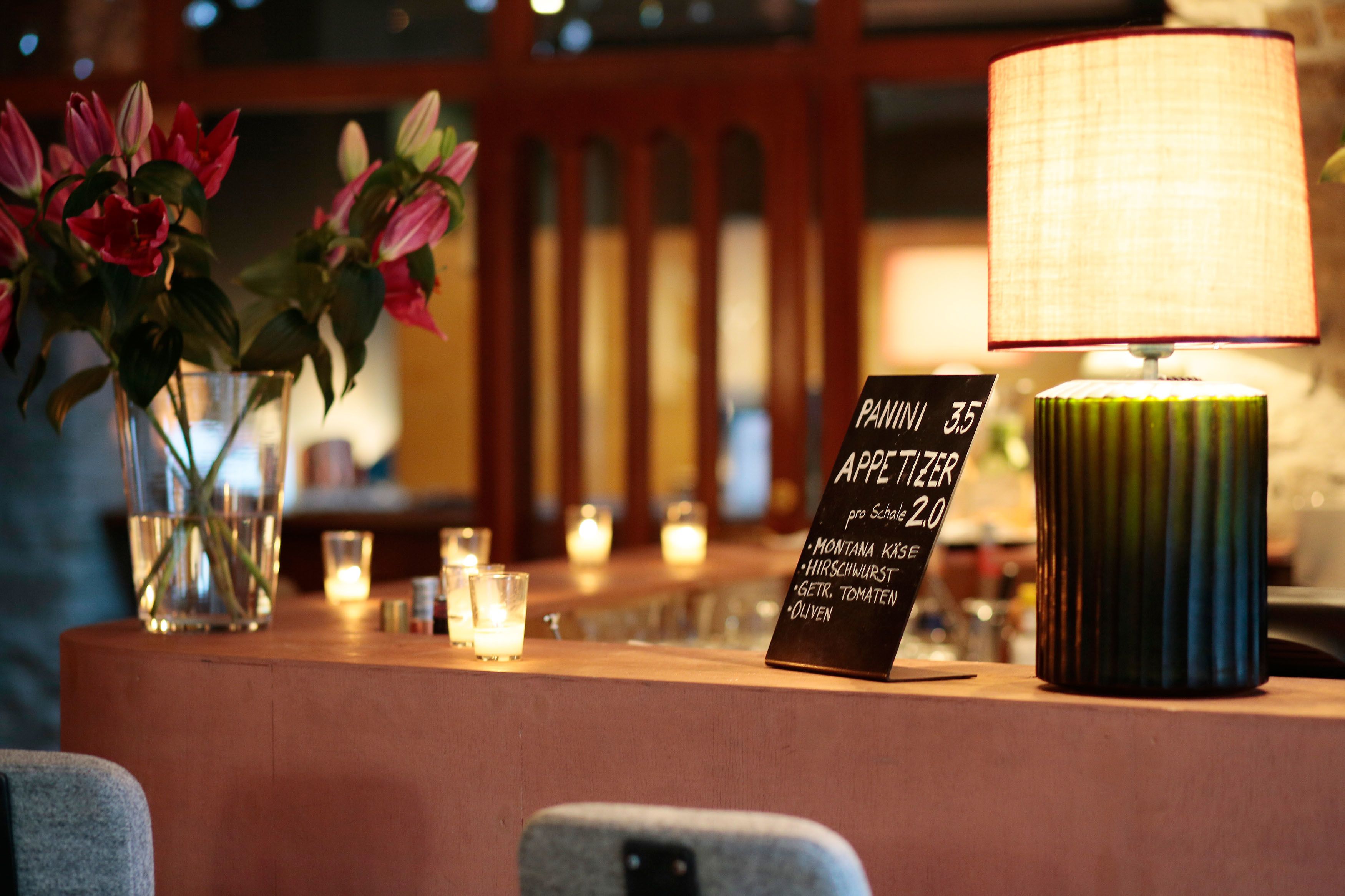 A bar area with a sign listing available drinks, a vase with flowers, and a lamp.