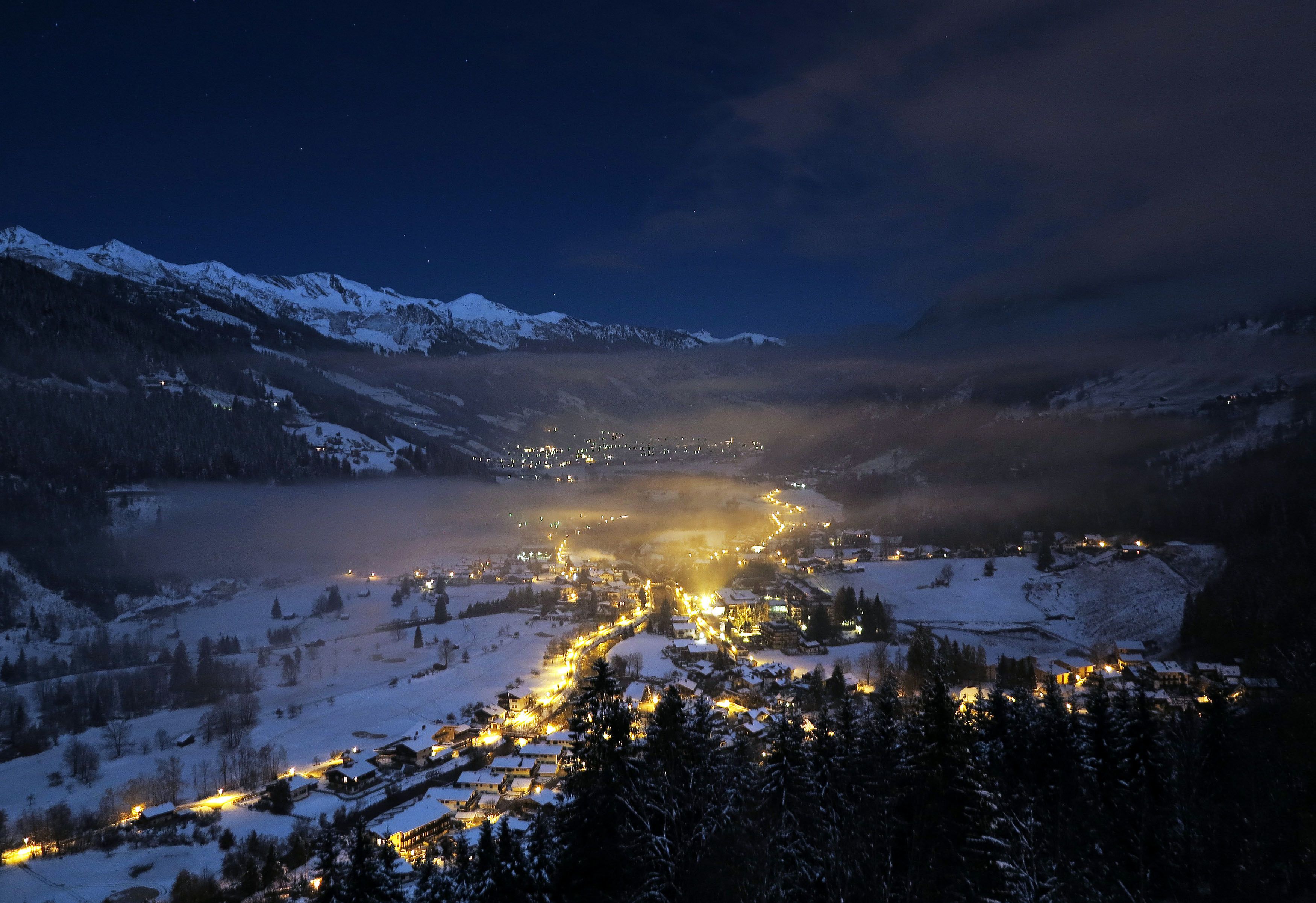Un villaggio montano innevato di notte, con edifici illuminati e una strada tortuosa.