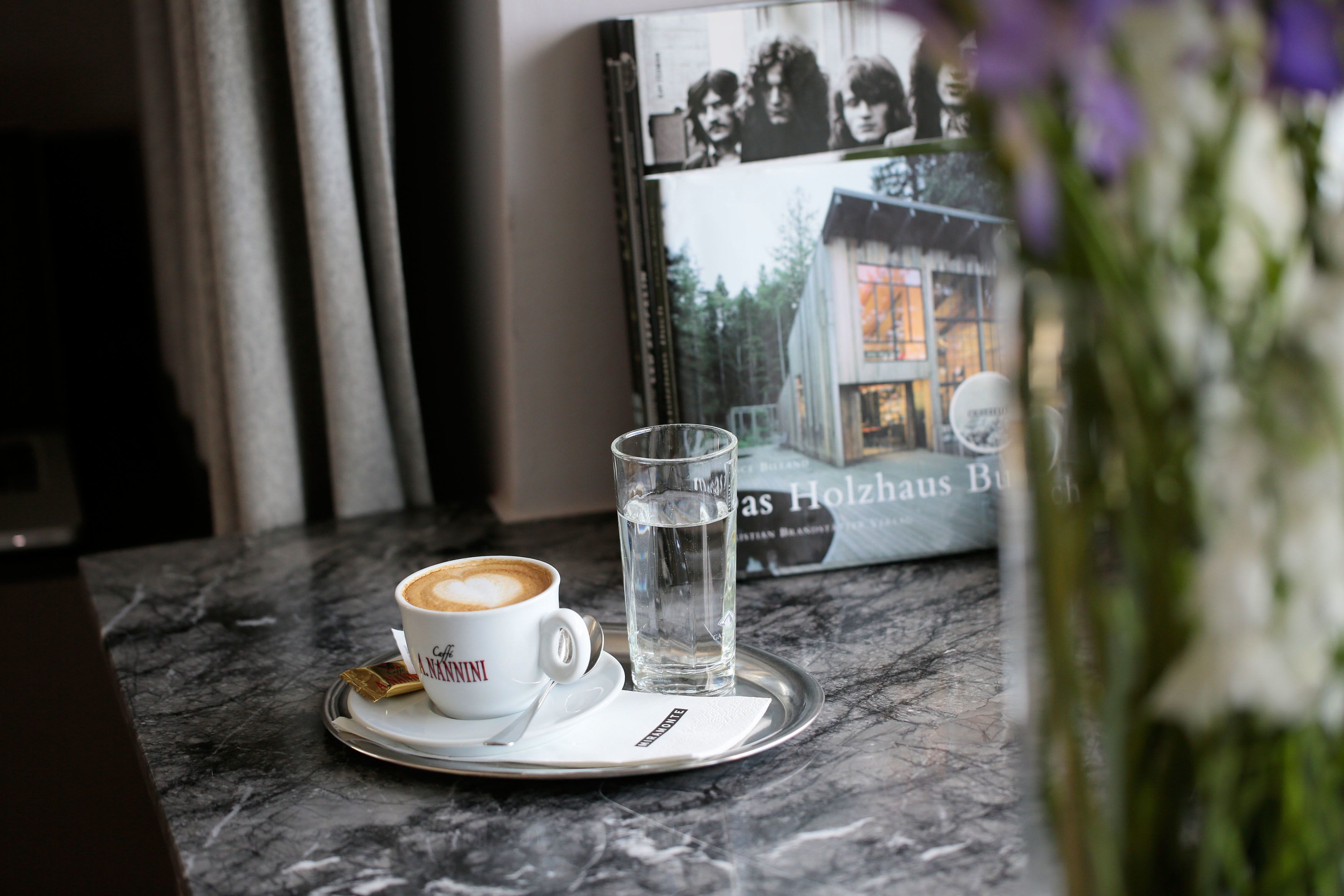 A coffee cup and glass of water on a tray, a book titled "As Holzhaus Buch," and a vase with flowers on a marble table.