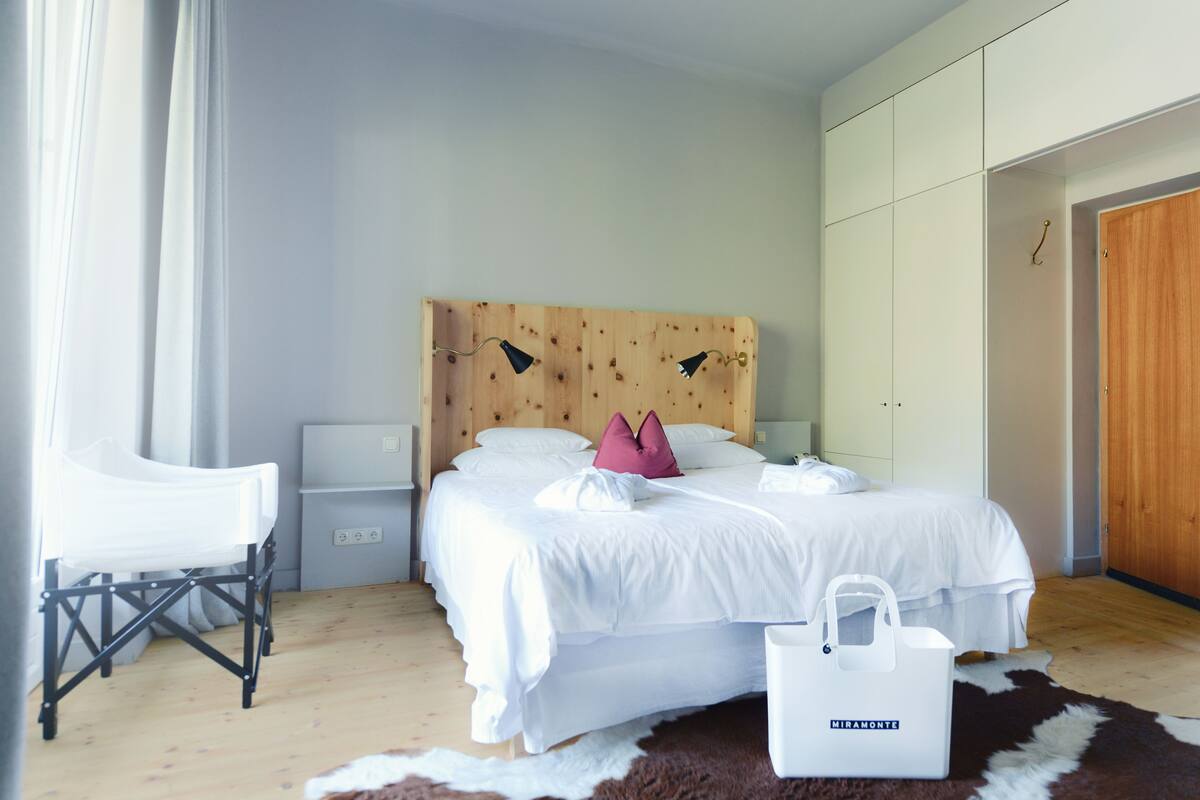 A bedroom with a wooden headboard, white bedding, a white chair, a small bedside table, a nightstand, a cowhide rug, and a white bag.