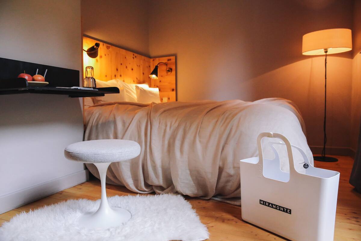 A bedroom with a bed, a white fluffy rug, a small white stool, a bedside table with a lamp, a wooden headboard, and a white bag on the floor.