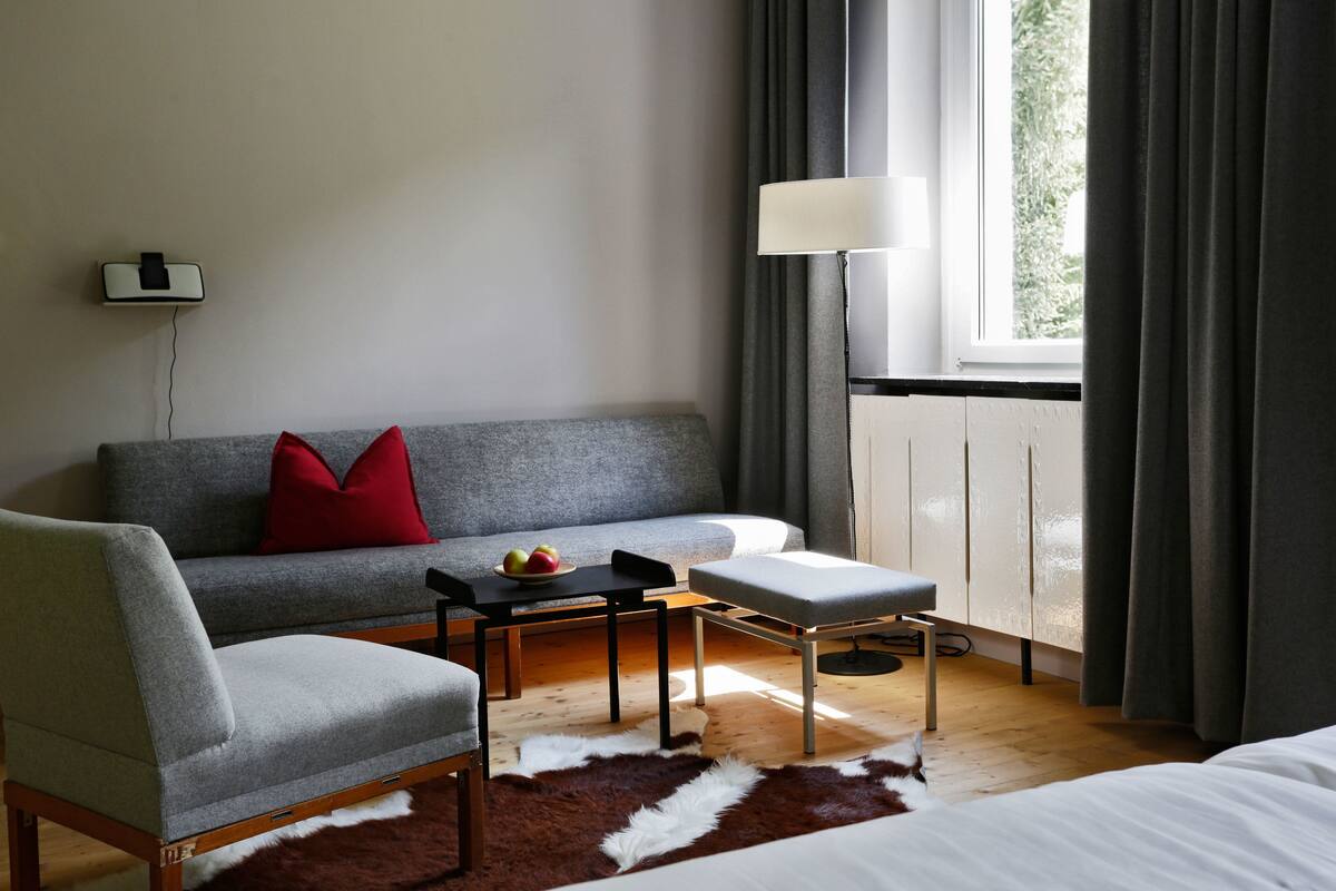 A modern hotel room with a grey sofa, a wooden chair, a small table with fruit, a white bed, and a large window with curtains.