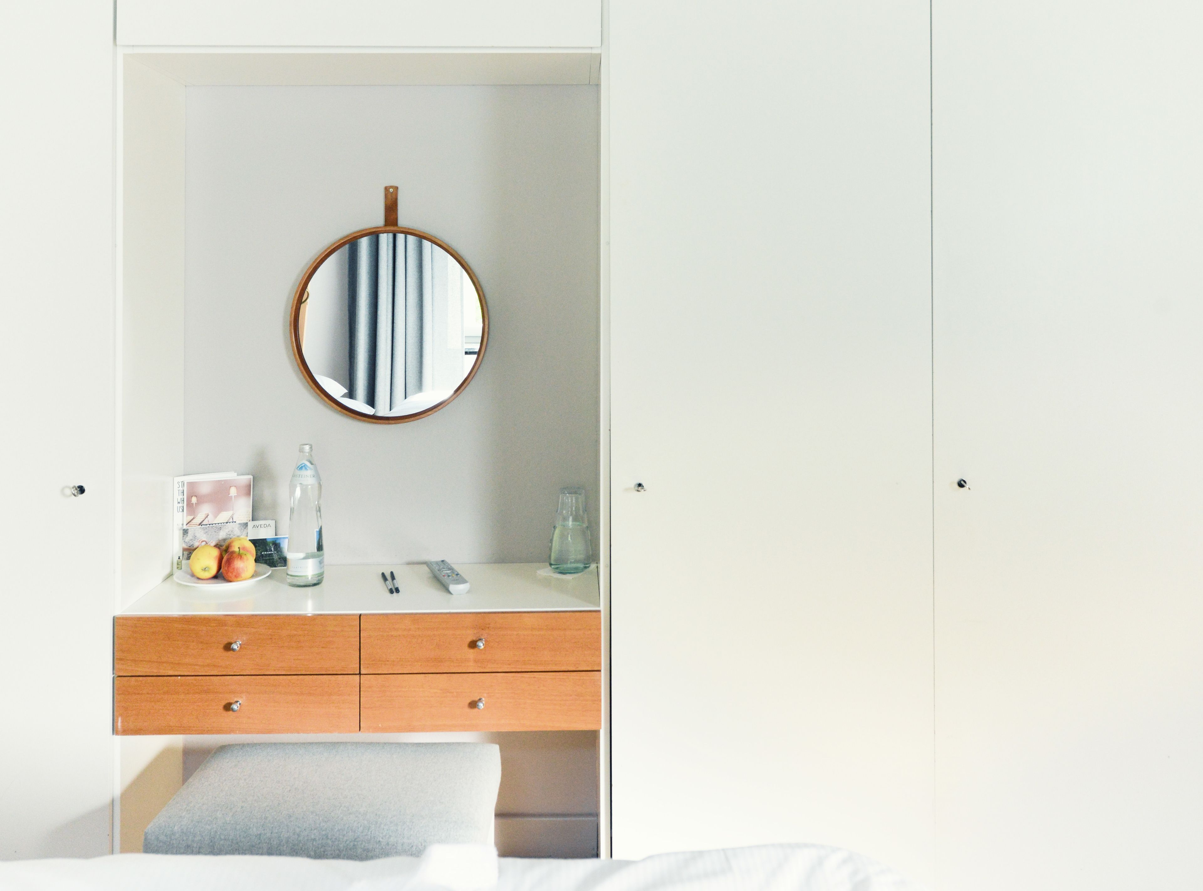 A white wardrobe with a round mirror, a wooden drawer, and a bedside table with a bottle, a remote control, and a glass.