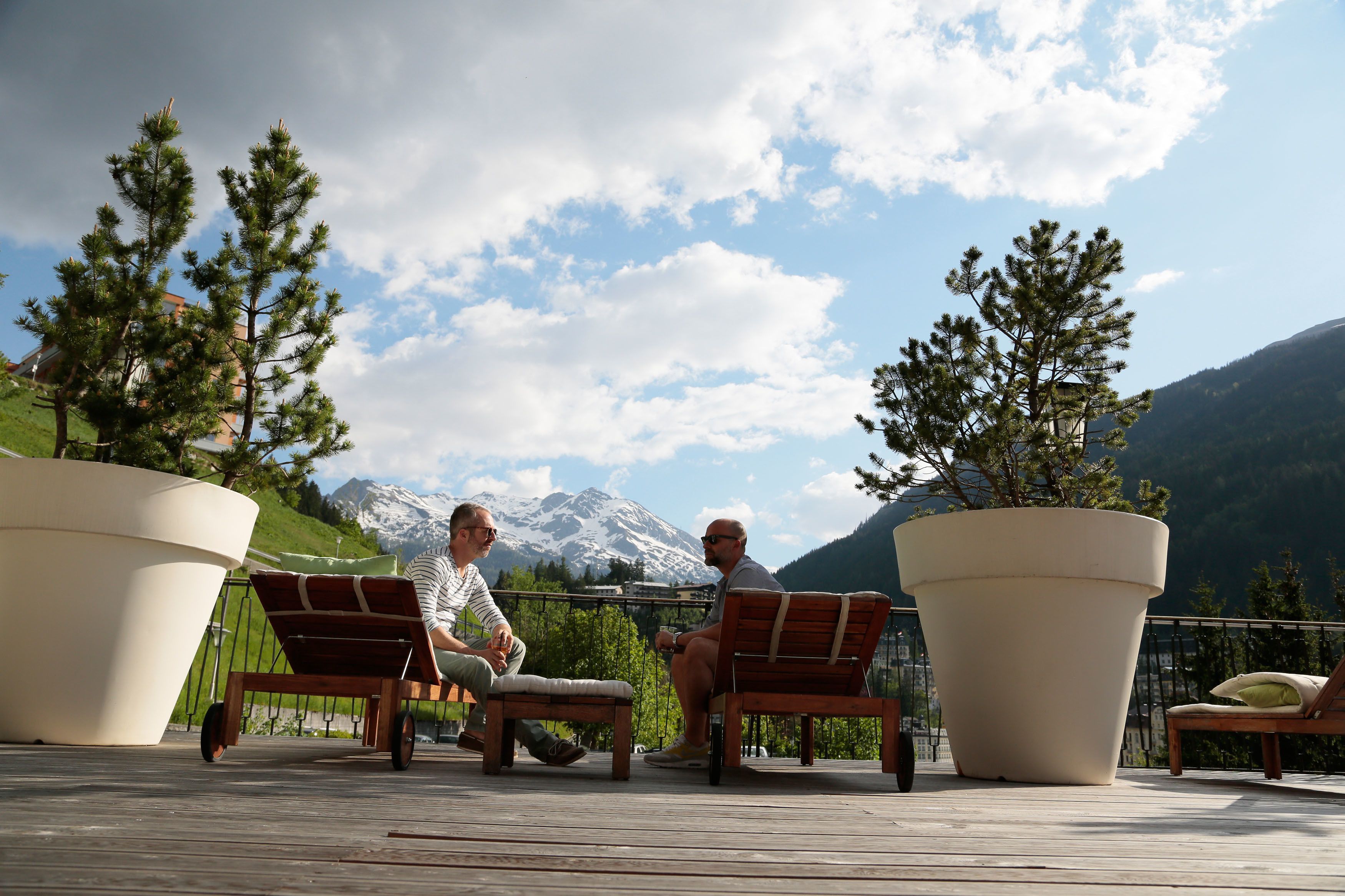 Two people seated on wooden chairs with large potted plants on a wooden deck, overlooking a mountainous landscape.
