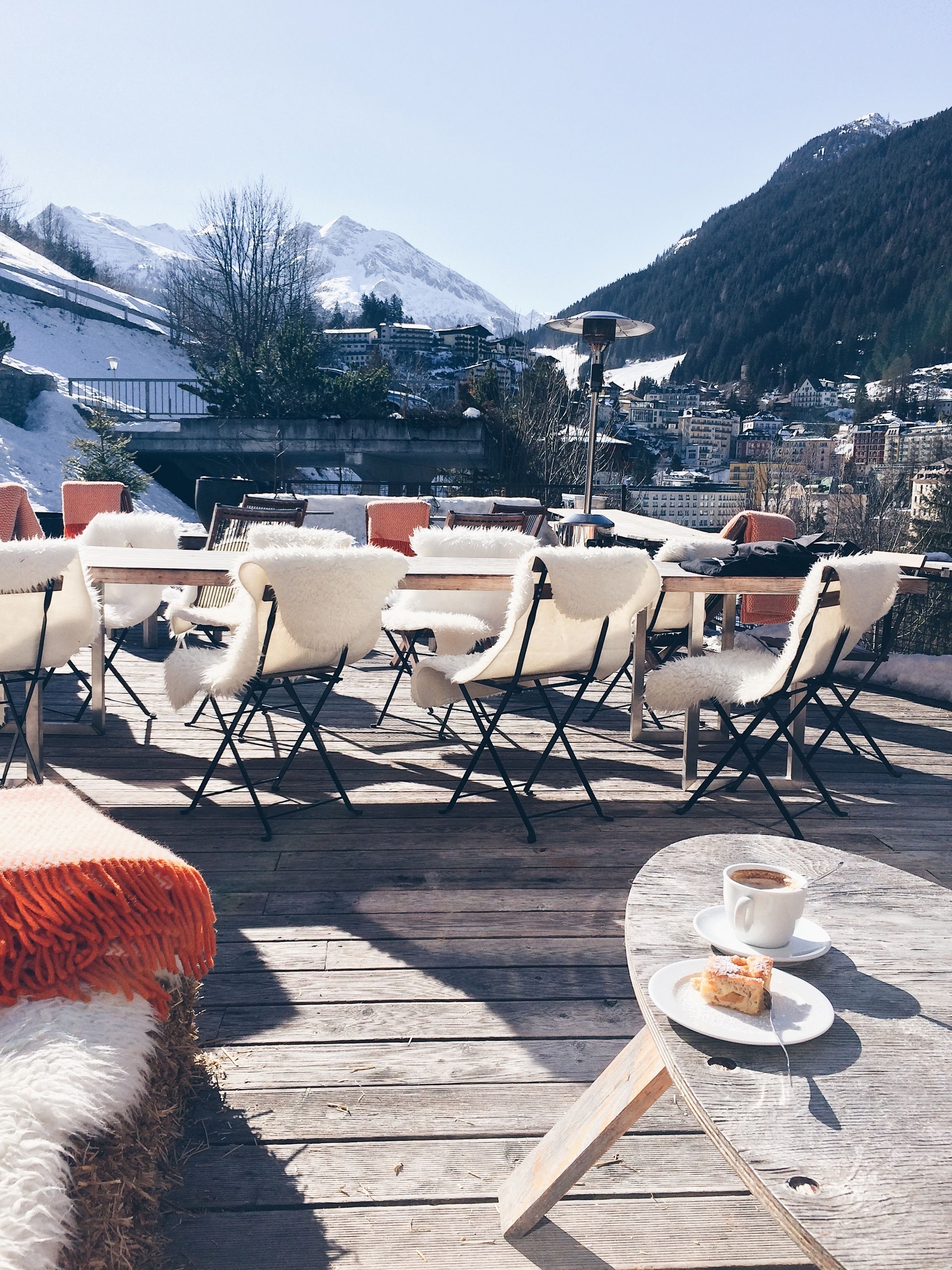 A wooden deck with white chairs and a table, a cup of coffee, and a slice of cake on the table.