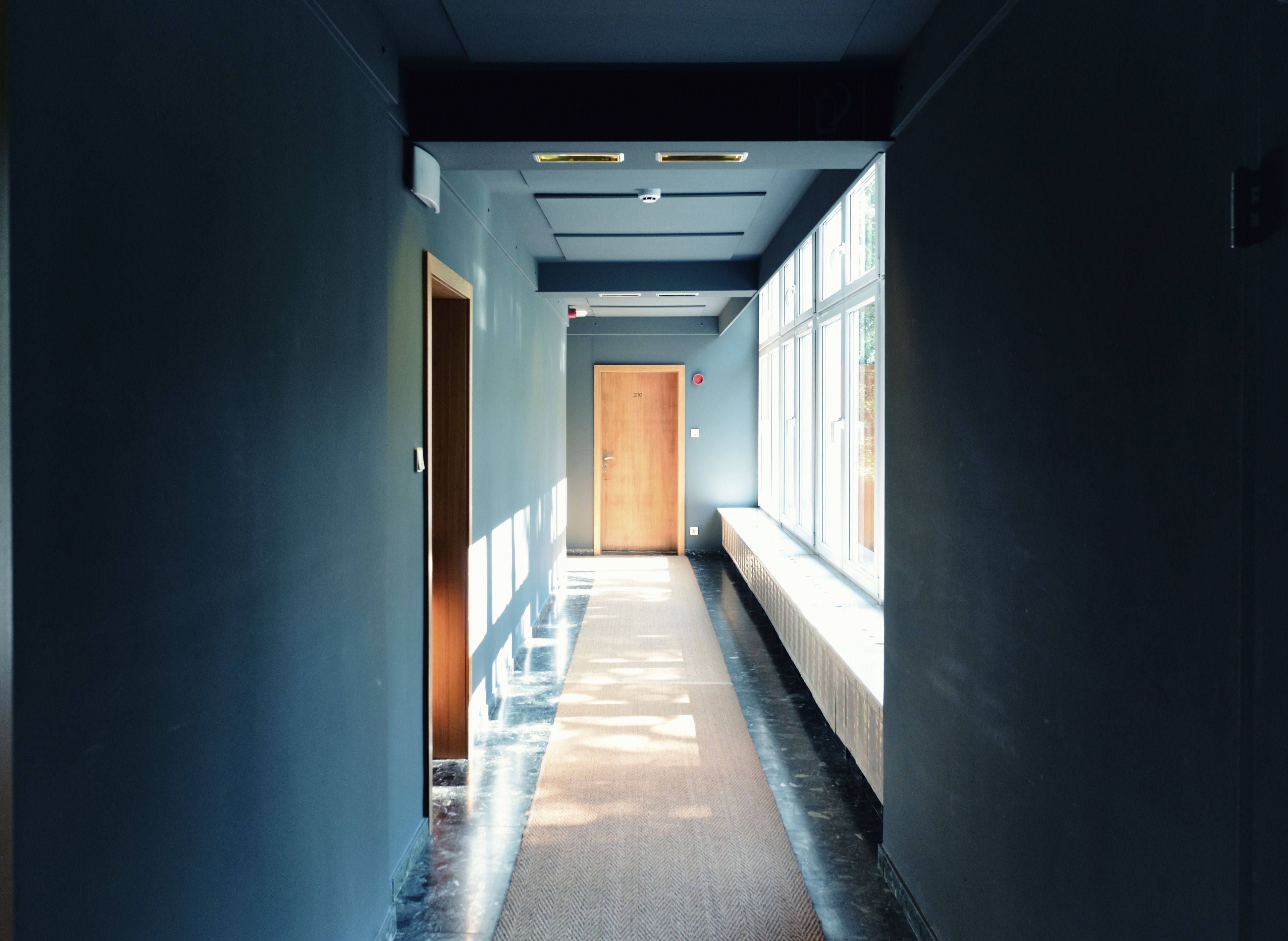 A long, narrow hallway with a wooden door at the end, large windows on one side, and a carpeted floor.