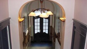 Een gang met een donkere vloer, een zwarte deur aan het einde, en twee lampen aan de muur.