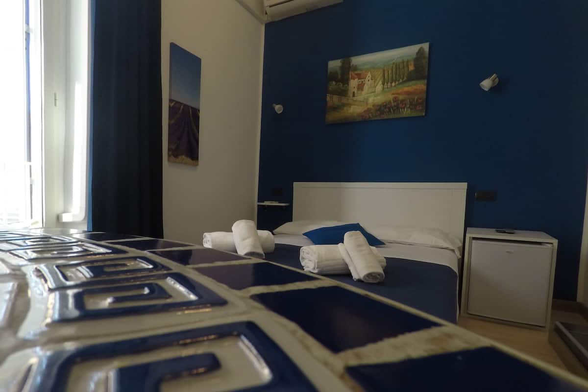 A hotel room with a bed, a small bedside table, a nightstand, and a painting on the wall.