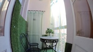 Une petite pièce avec une table ronde et deux chaises, une plante en pot et une vue sur l’extérieur à travers une fenêtre ornée de rideaux légers.